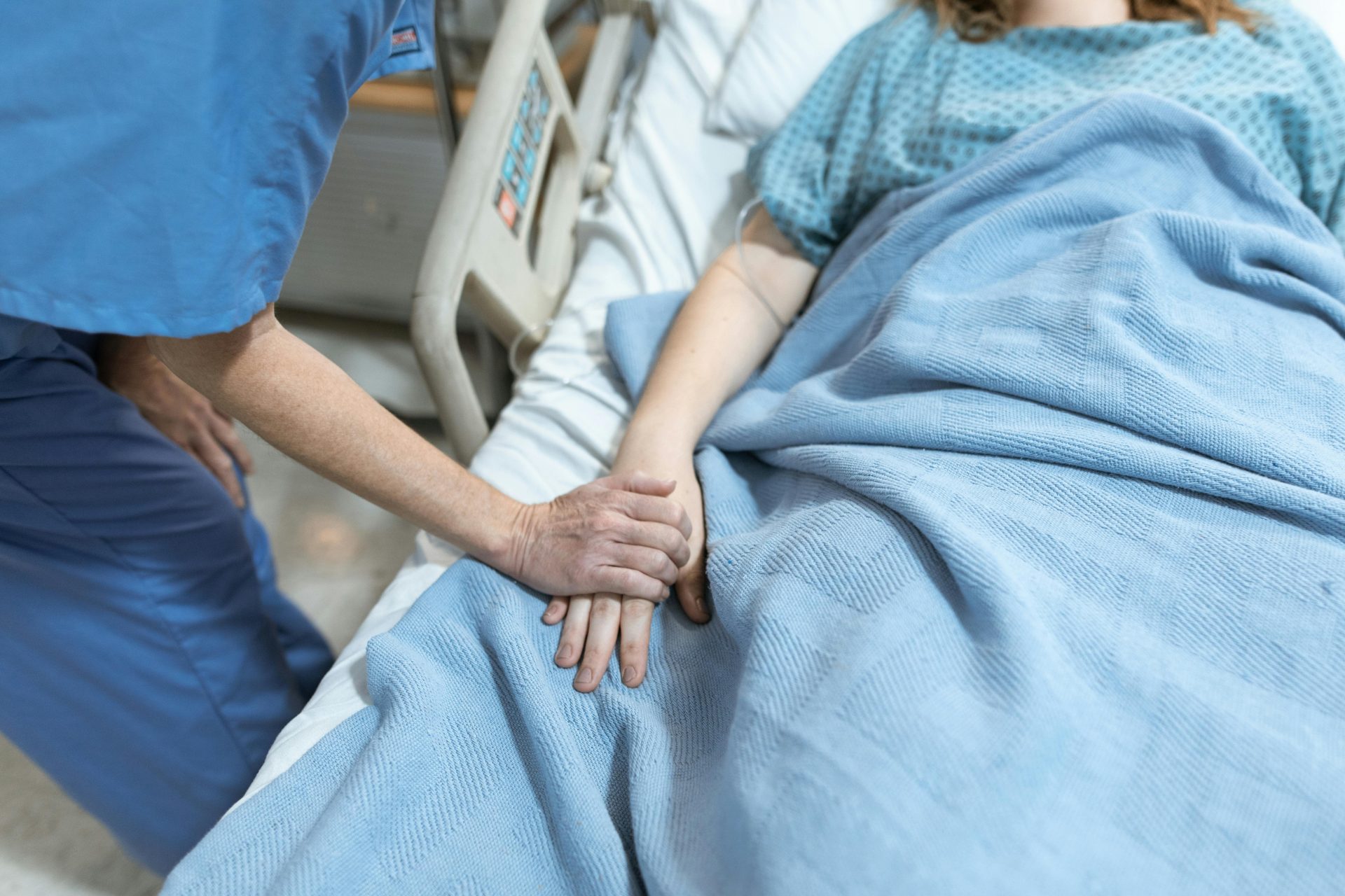 Eine Krankenschwester im OP-Kittel spendet Trost, indem sie einem Patienten im Krankenhausbett die Hand hält.