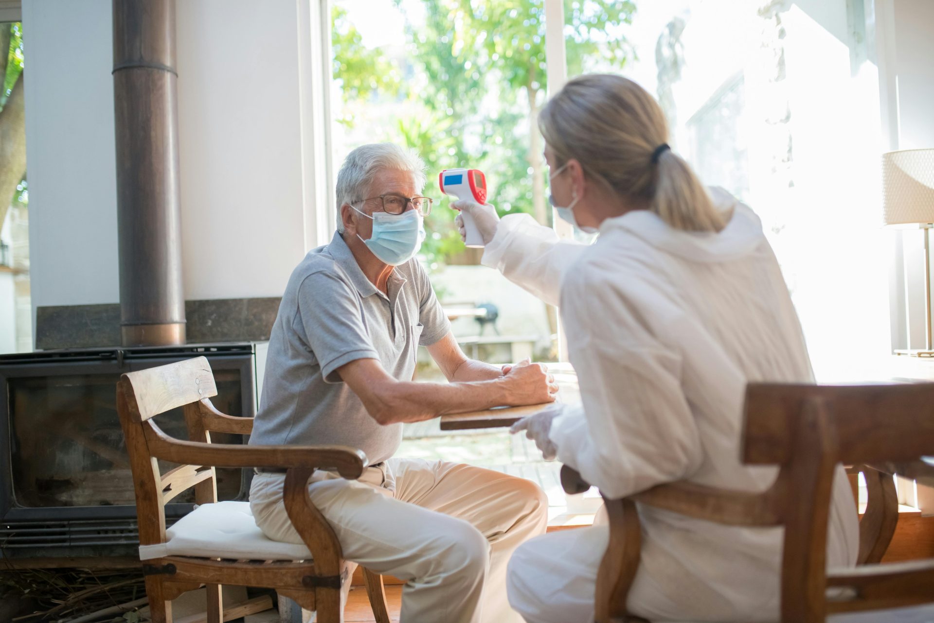 Ein älterer Mann in Schutzanzug und Maske wird im Innenbereich von einem Mitarbeiter des Gesundheitswesens einer Temperaturkontrolle unterzogen.